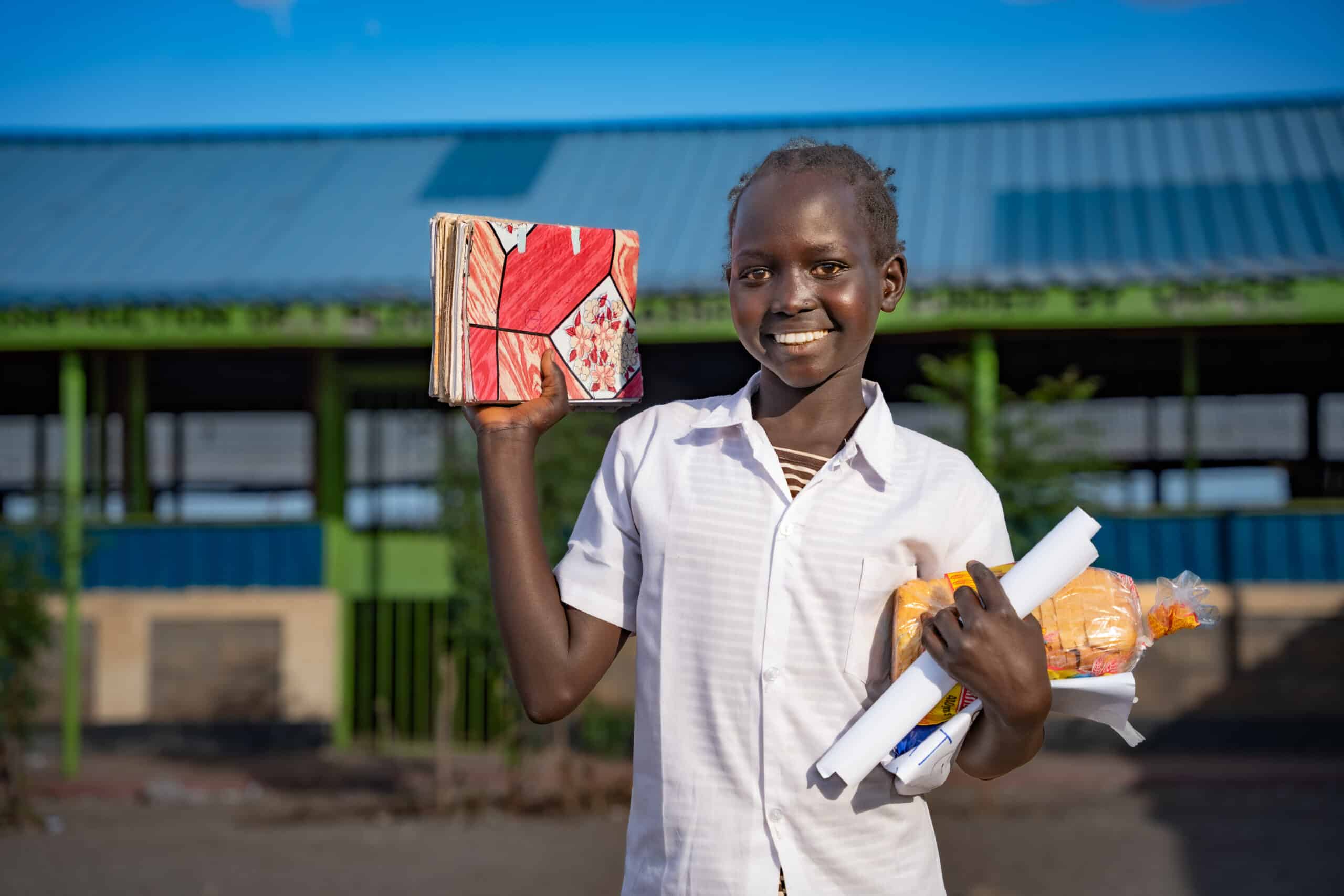 A young student holding books smiles outside of her classroom in Kenya.